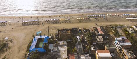 An aerial view of a coastal area with a beach, beach umbrellas, and a cluster of buildings with blue roofs.