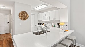 A modern kitchen with white cabinetry, a central island with a sink, and a decorative wall piece. - First Floor Condo • Walk to Beach • Sleeps 4 (Mexico Beach)