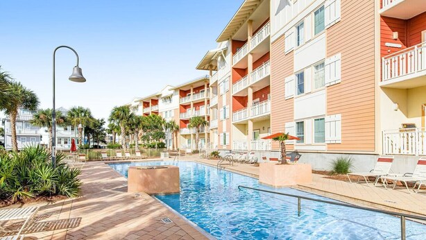 A swimming pool area with a clear view of a multi-story building with balconies, a street lamp, and a palm tree. - First Floor Condo • Walk to Beach • Sleeps 4 (Mexico Beach)