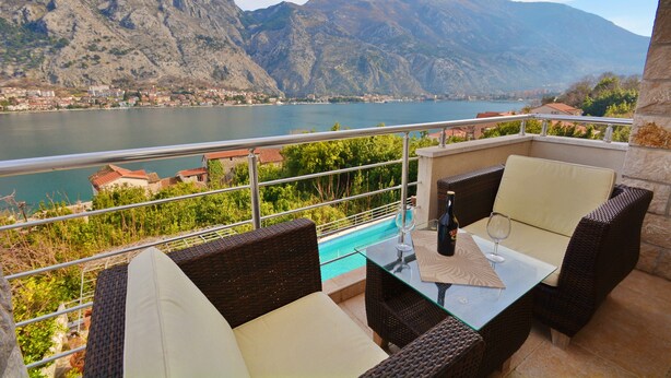 A balcony with wicker furniture, a glass table, and a view of a lake and mountains. - Muo Apartments (Kotor)