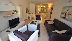 A modern living room with a flat-screen TV, white and black furniture, a dining area with a table and chairs, and a kitchen in the background. - Muo Apartments (Kotor)