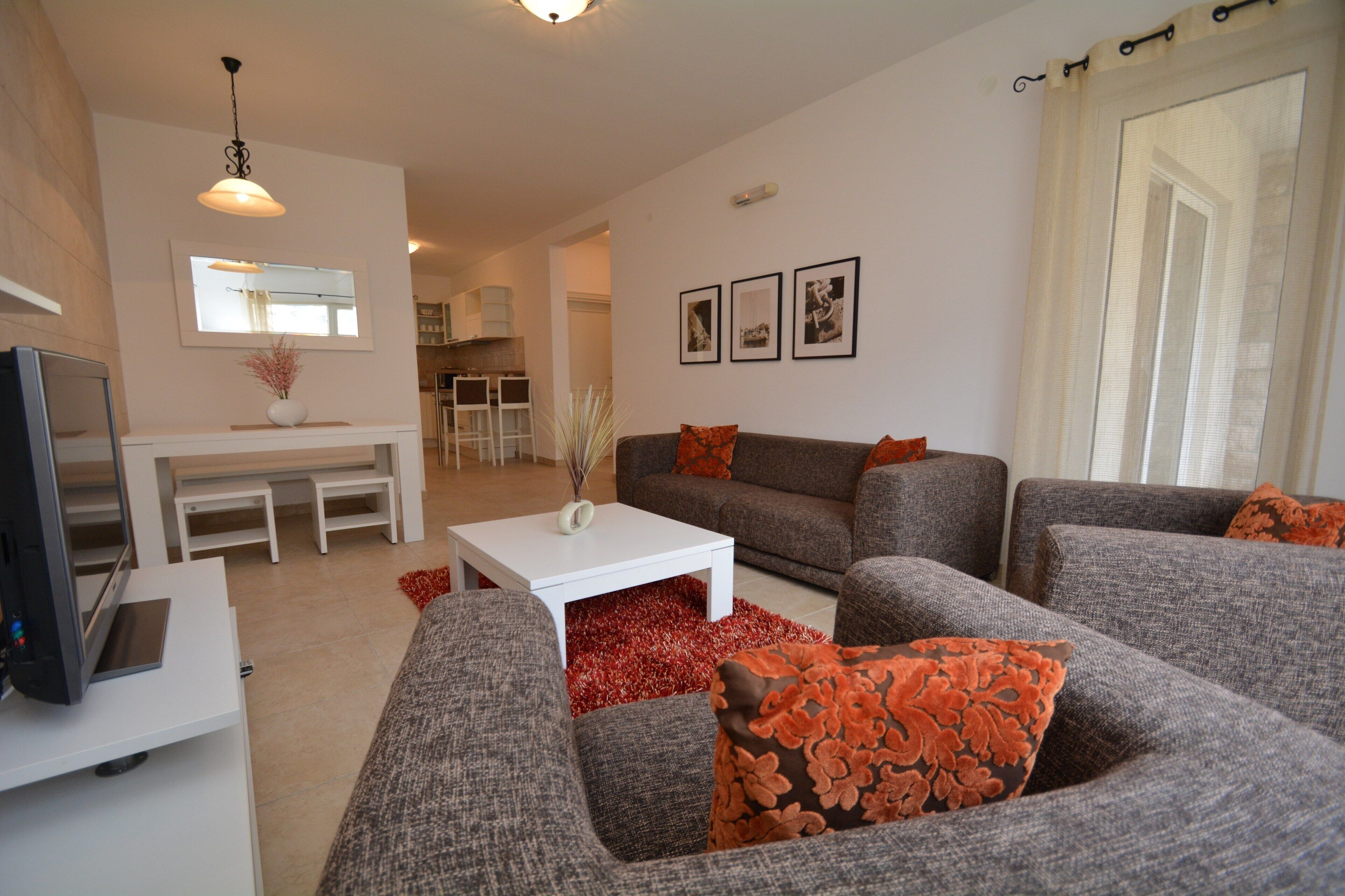 A modern living room with a grey sofa set, a white coffee table, and a TV on a white stand. There is a dining area with a white table and chairs in the background.