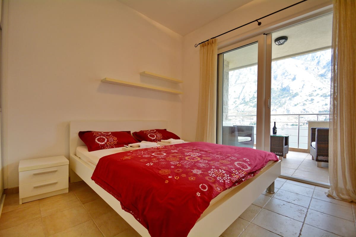 A bedroom with a bed, a nightstand, a window with curtains, and a view of a balcony.