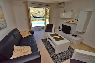 Una sala de estar moderna con un sofá de cuero negro, una mesa de centro blanca y una televisión. Se ve agua y un barco al fondo.