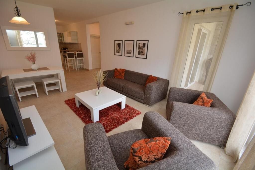 Ein Wohnzimmer mit grauem Sofa, einem weißen Couchtisch und einem Fernseher auf einem Schrank. Auf dem Boden liegt ein roter Teppich, und im Hintergrund ist eine Küche erkennbar.