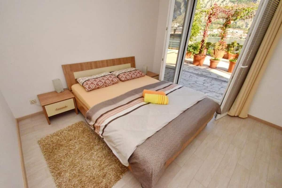 A bedroom with a bed, a nightstand, a small drawer, a window with a view of potted plants and a building.