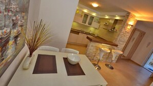 A dining area with a table, chairs, and a vase of dried plants. There is a view of a kitchen and a dining area in the background. - Muo Apartments (Kotor)