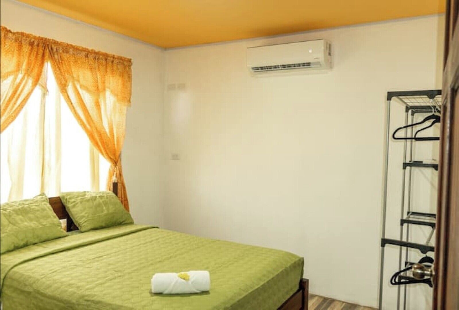 A single bed with a green bedspread and pillows, an air conditioning unit on the wall, and a clothing rack with hangers.