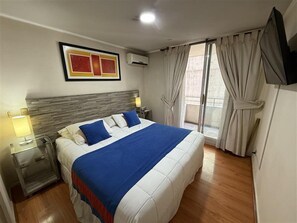 A hotel room with a large bed, a headboard, two bedside tables with lamps, a flat-screen TV, and a window with curtains. - VR Suite (Santiago)