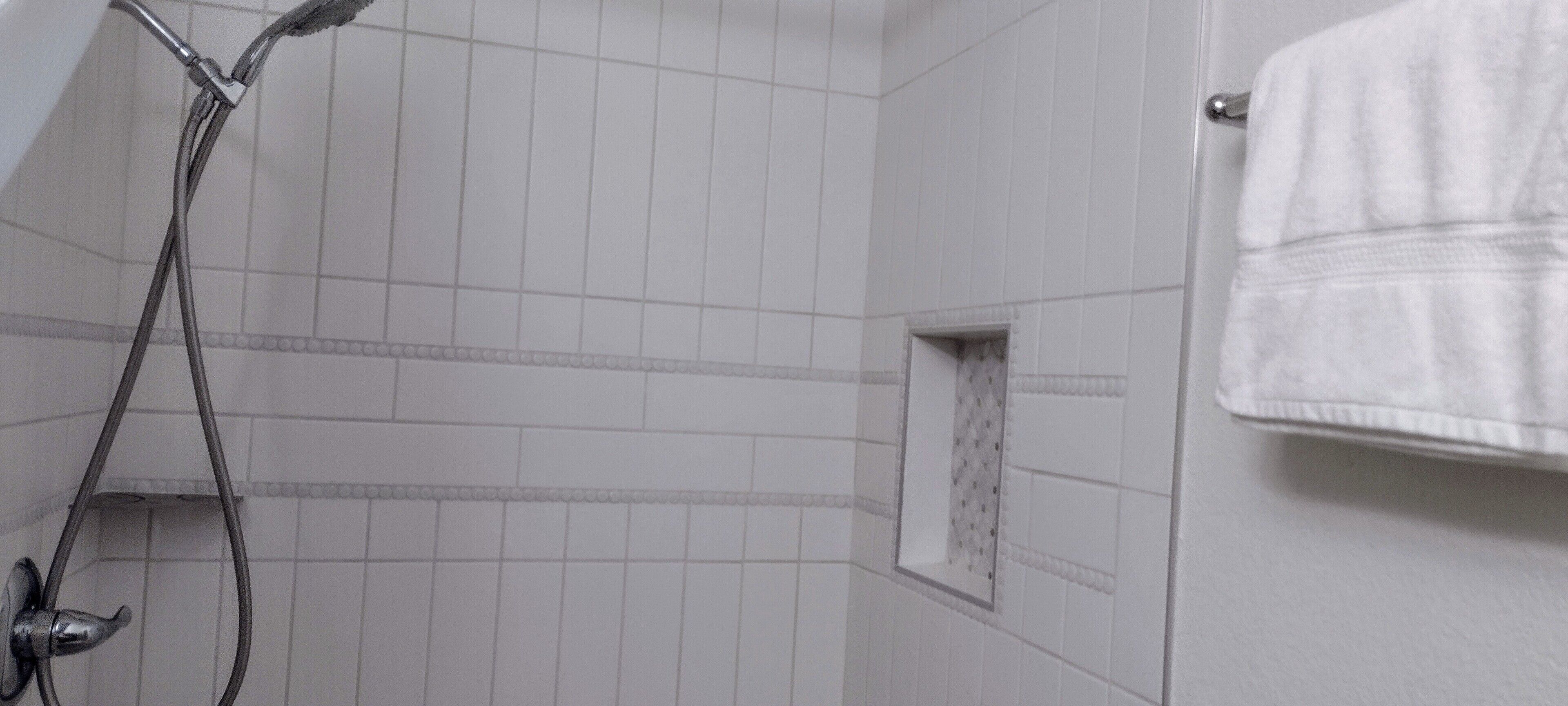 Un baño con paredes de azulejos blancos, ducha con cabezal de mano y un toallero con una toalla blanca.