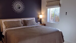 A neatly made bed with a quilted cover, two bedside lamps, a wall clock, and a window with a view of greenery.