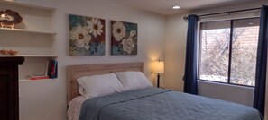 Une chambre à coucher avec un lit, une table de chevet avec une lampe, deux tableaux représentant des motifs floraux accrochés au mur et une fenêtre donnant sur des arbres.