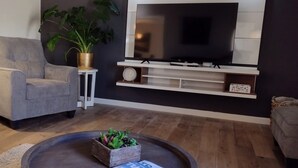 A modern living room with a grey sofa, a small side table with a plant, a wooden coffee table with a plant, a TV on a wall-mounted shelf, and a clock on the side table.