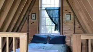 A cozy attic bedroom with a wooden ceiling, a large window with a sheer curtain, and a bed with blue bedding.