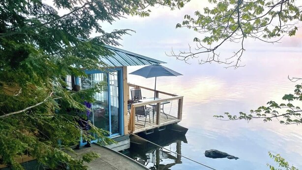 A floating house with a glass door, a deck with outdoor furniture, and a view of the water.