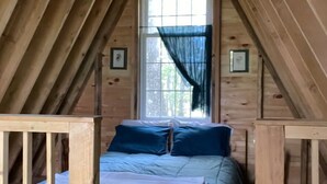 Een gezellig slaapkamer op zolder met een houten plafond, een groot raam met dunne gordijnen en een bed met blauw beddengoed.