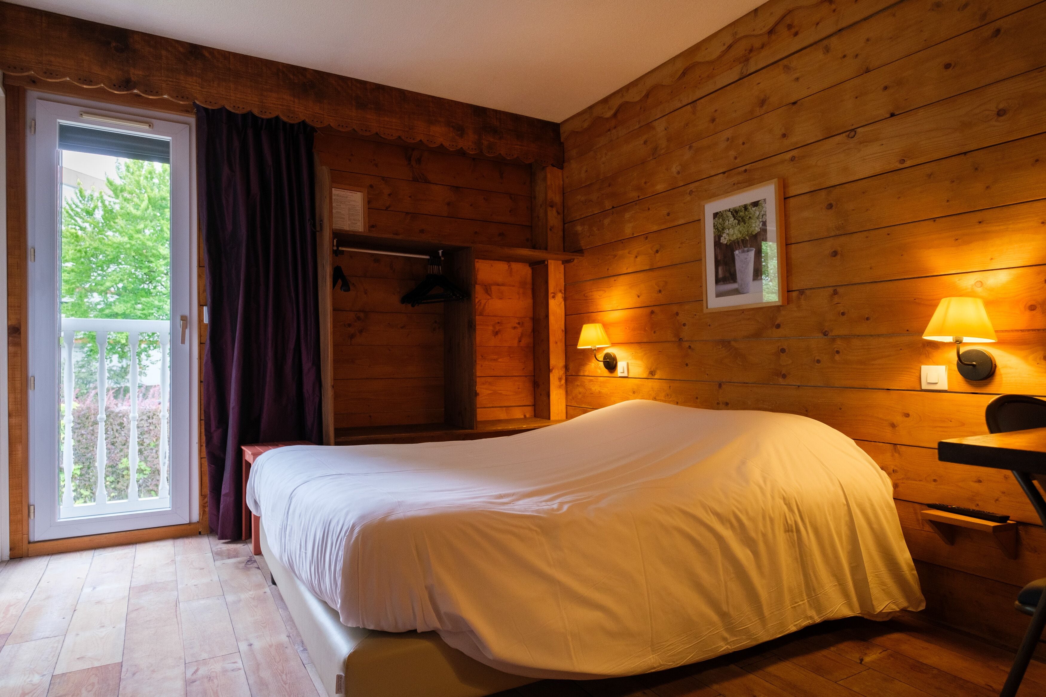 A wooden cabin room with a bed, a door leading to a balcony, and two wall-mounted lamps.