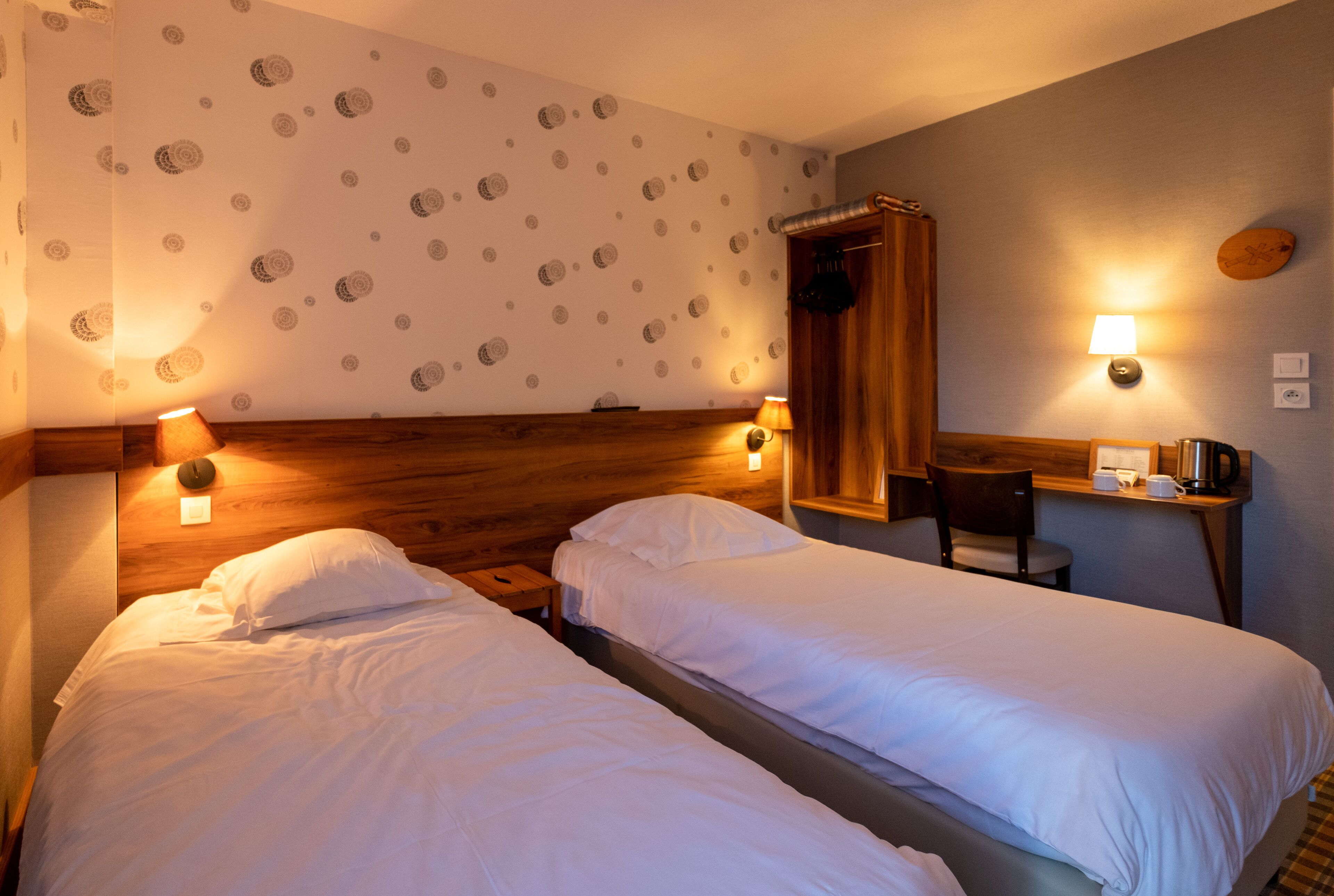 Habitación de hotel con dos camas individuales, cabecera de madera, un escritorio con silla, un espejo y dos lámparas de pared.