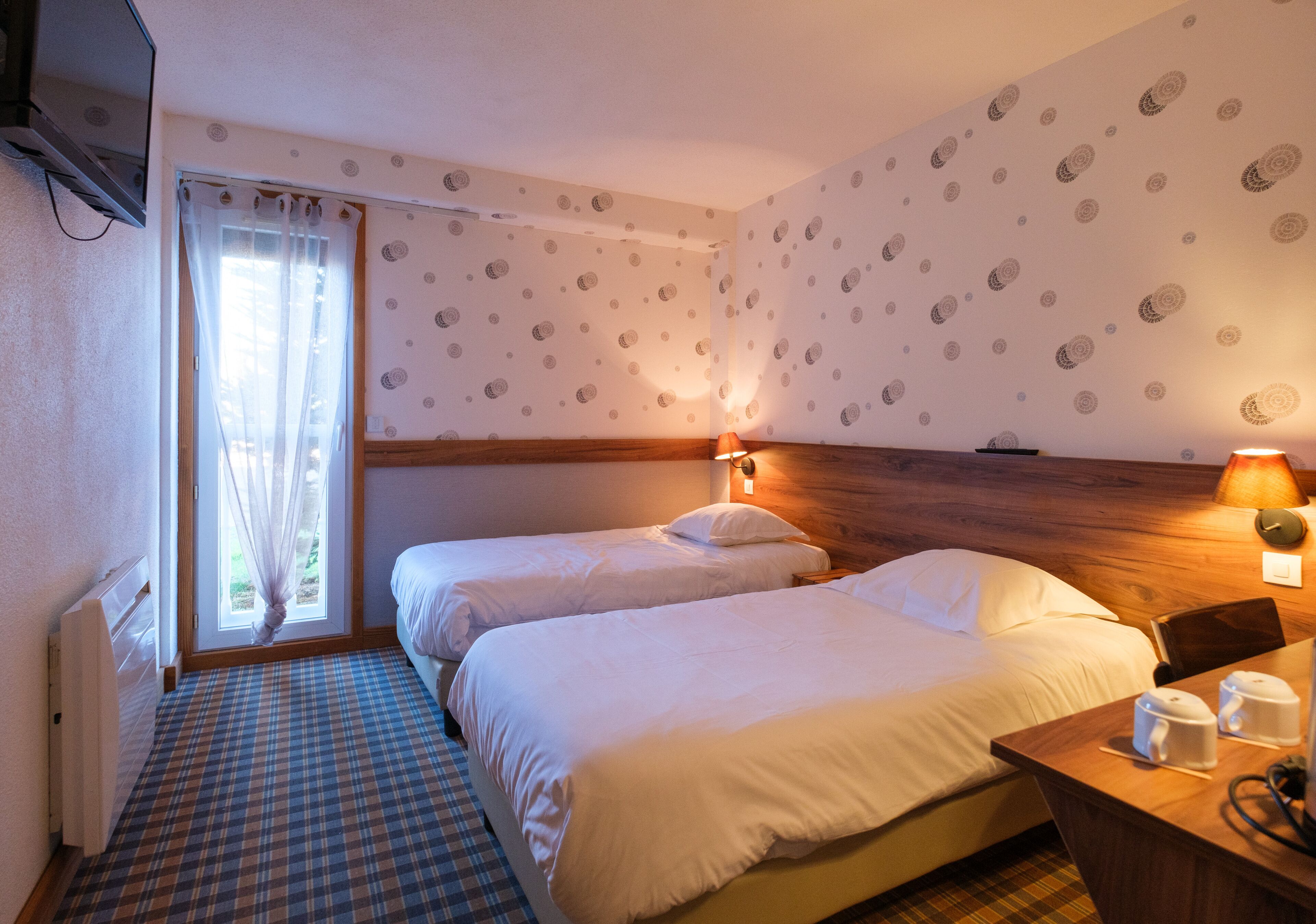 Habitación de hotel con dos camas, cabecero de madera, ventana con cortinas translúcidas, pared con diseño y mesita de noche con lámpara.