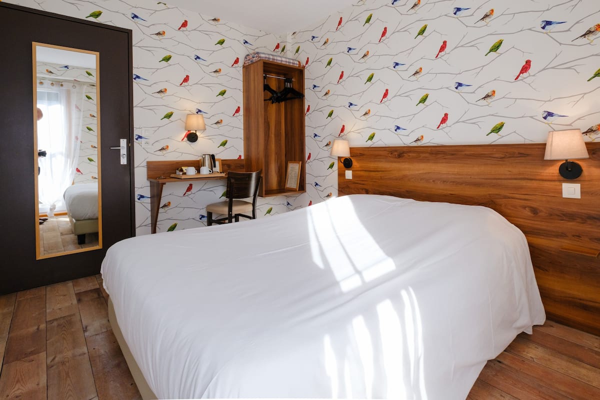 Ein Schlafzimmer mit hölzernem Kopfteil, einem Bett mit weißer Bettwäsche, einem Schreibtisch mit Stuhl, einem Spiegel und einer Wand mit Vogelmuster.