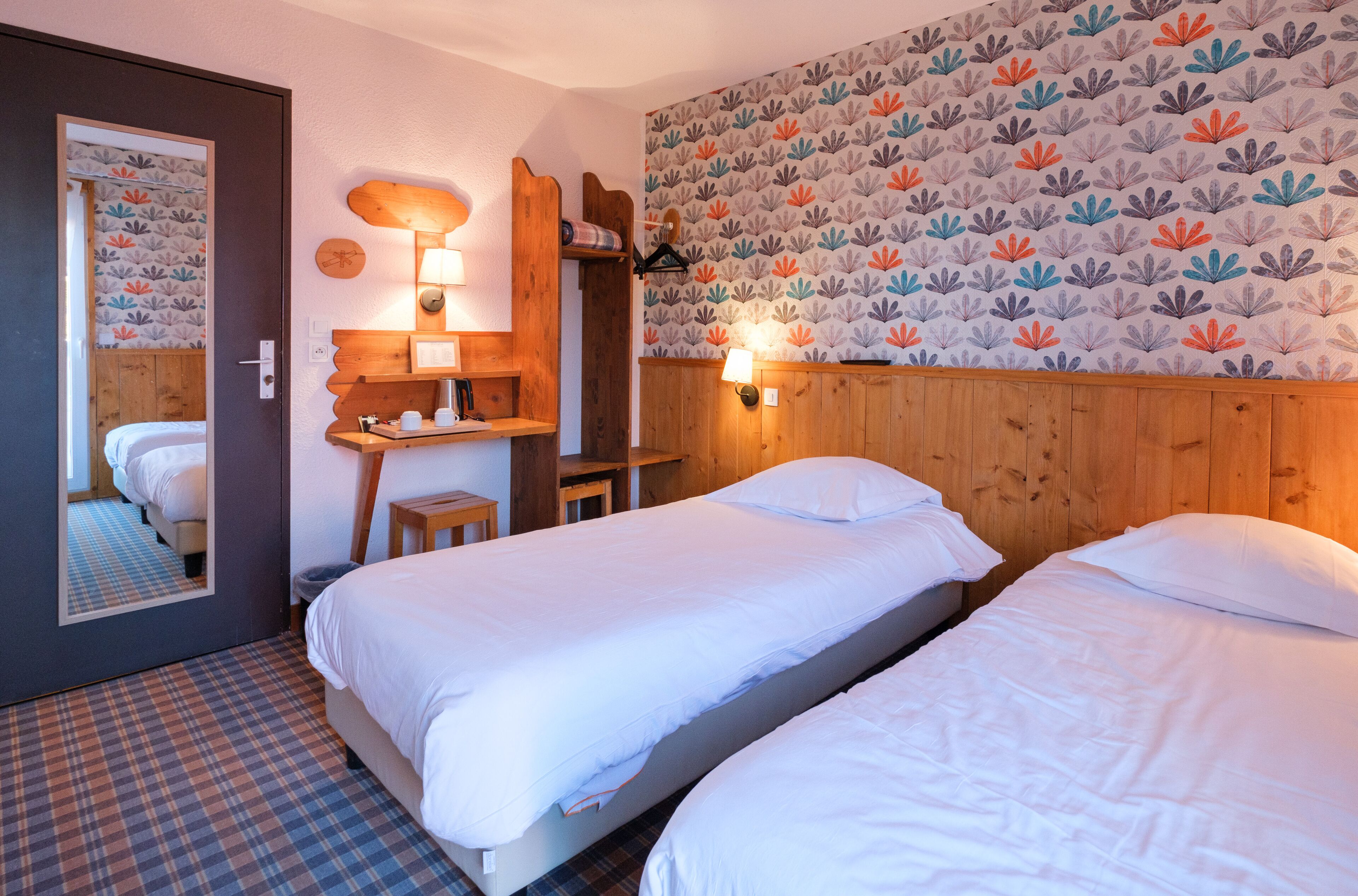 A hotel room with two beds, a wooden headboard, a mirror, a small desk with a lamp, and a patterned wallpaper.