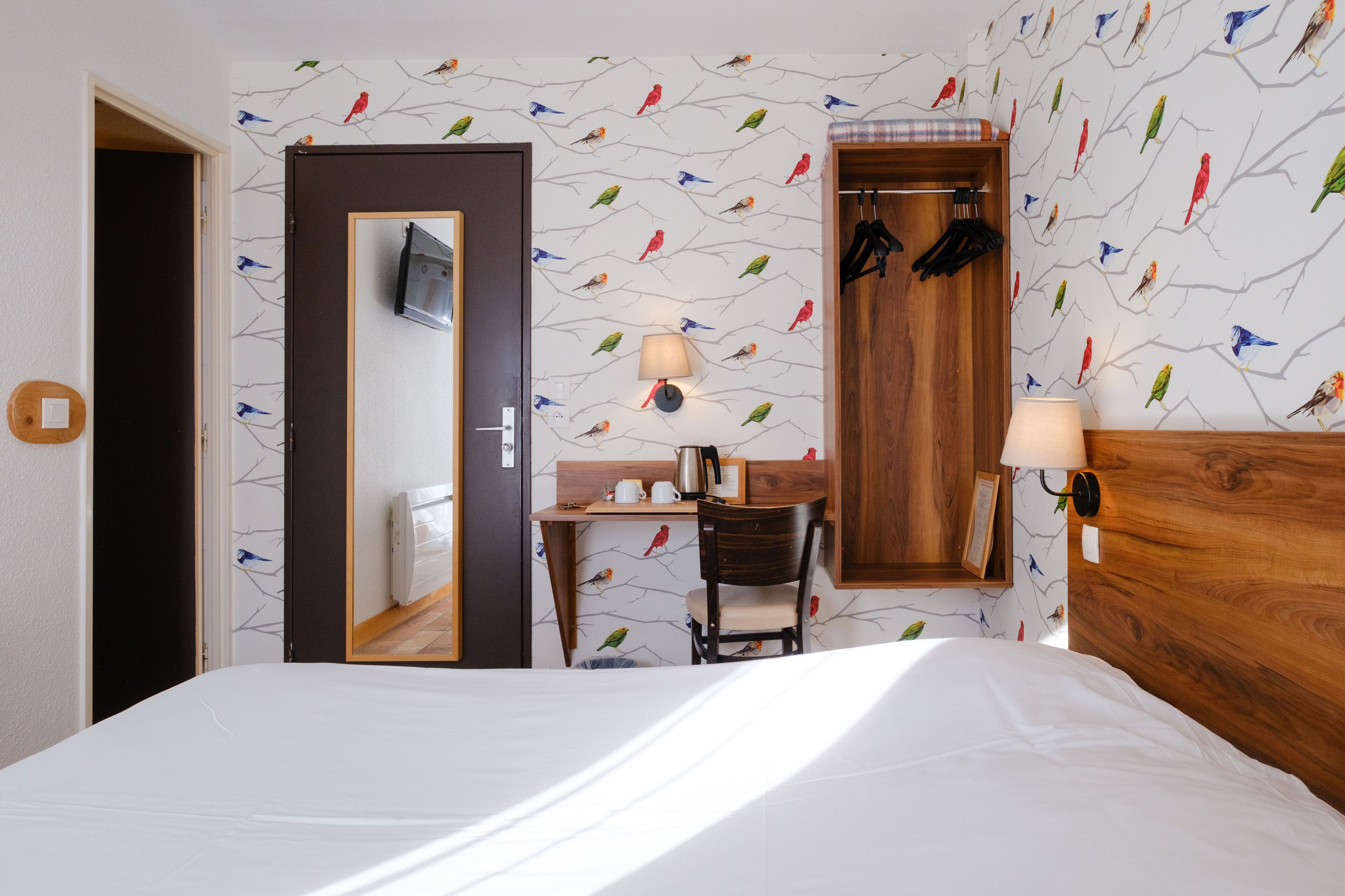 A hotel room with a bed, a desk with a chair, a wooden wardrobe, a wall-mounted TV, and a patterned wallpaper.