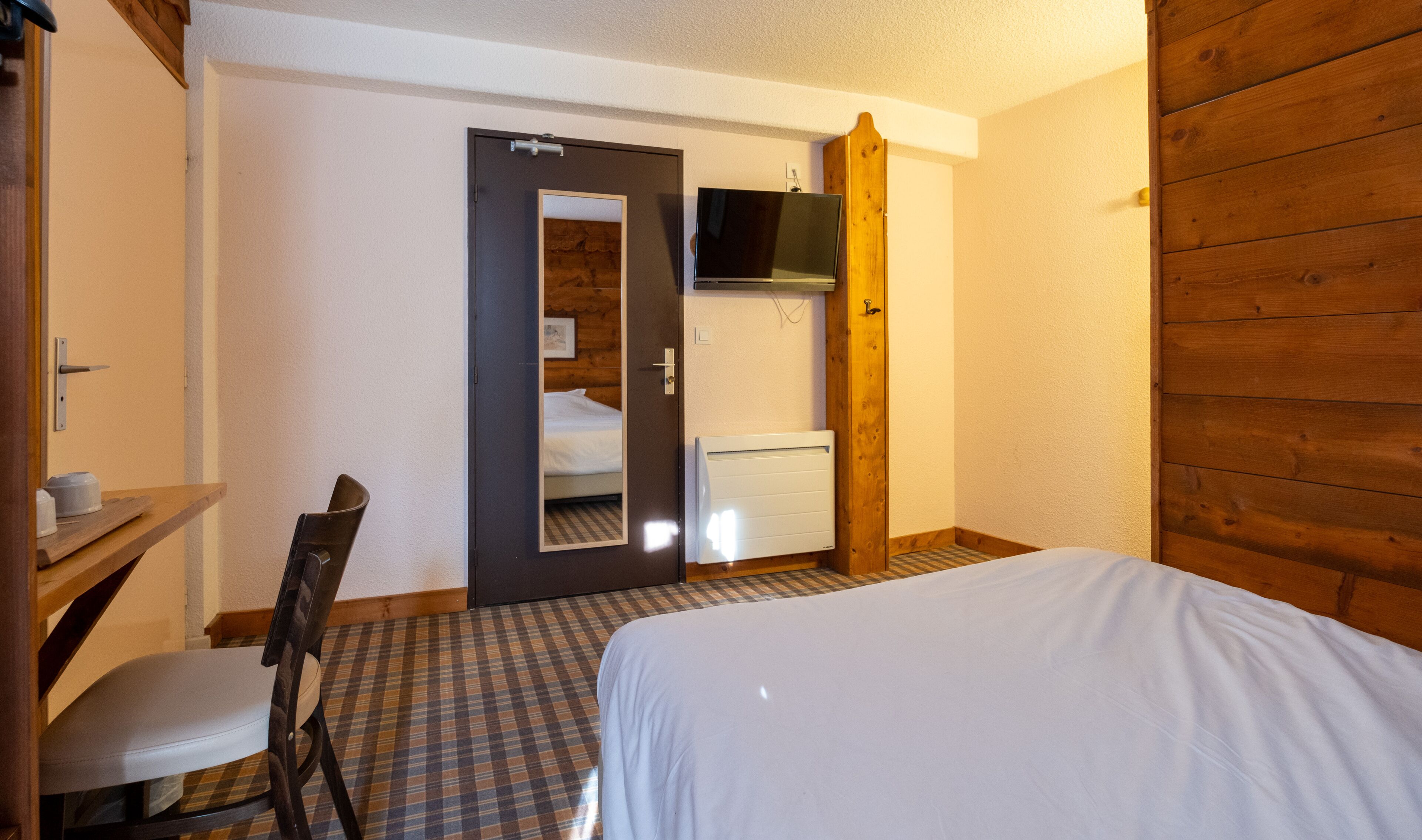 Una habitación pequeña y acogedora con cama, sillón, escritorio y televisor sobre un panel de madera.