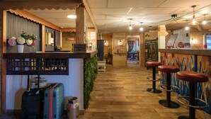 Een bar met houten vloer, een barbalie en twee krukken. Bij de ingang staan koffers en een decoratieve vaas.