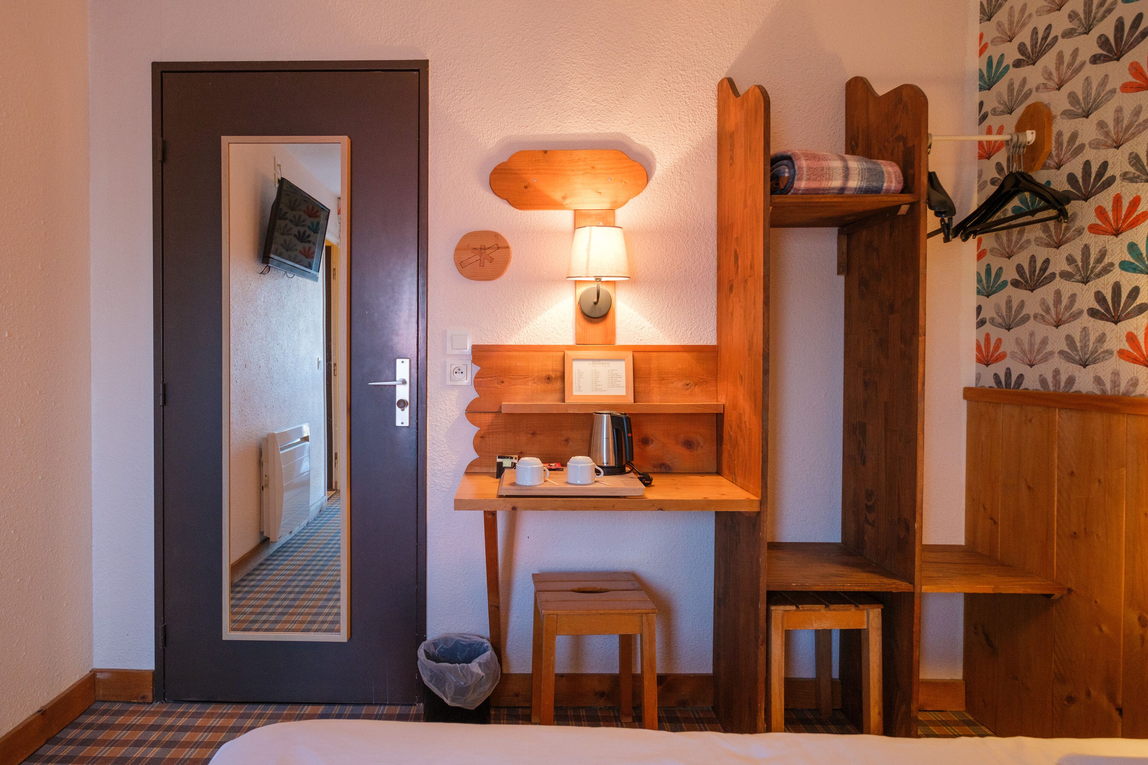 Habitación de hotel con un escritorio de madera, un taburete pequeño, un espejo, una lámpara de pared y un estante con una tetera y tazas.