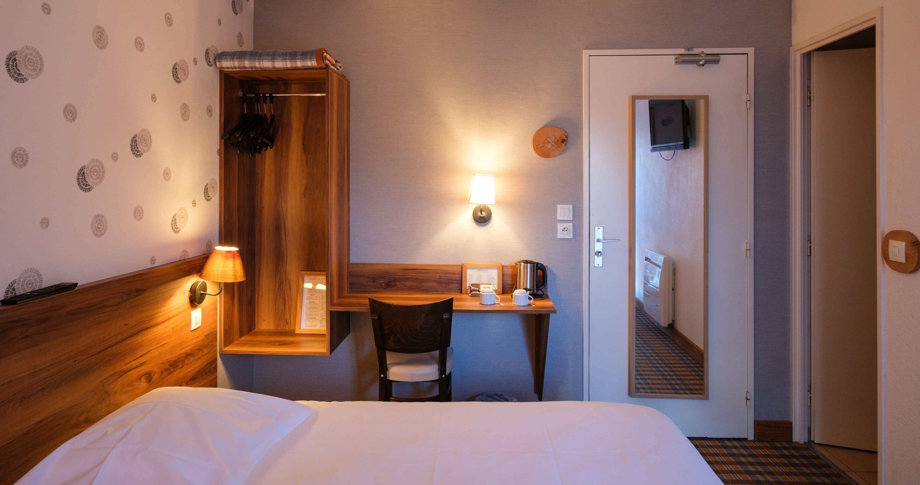 A hotel room with a bed, a desk with a chair, a mirror, a wall-mounted lamp, and a door leading to a hallway.