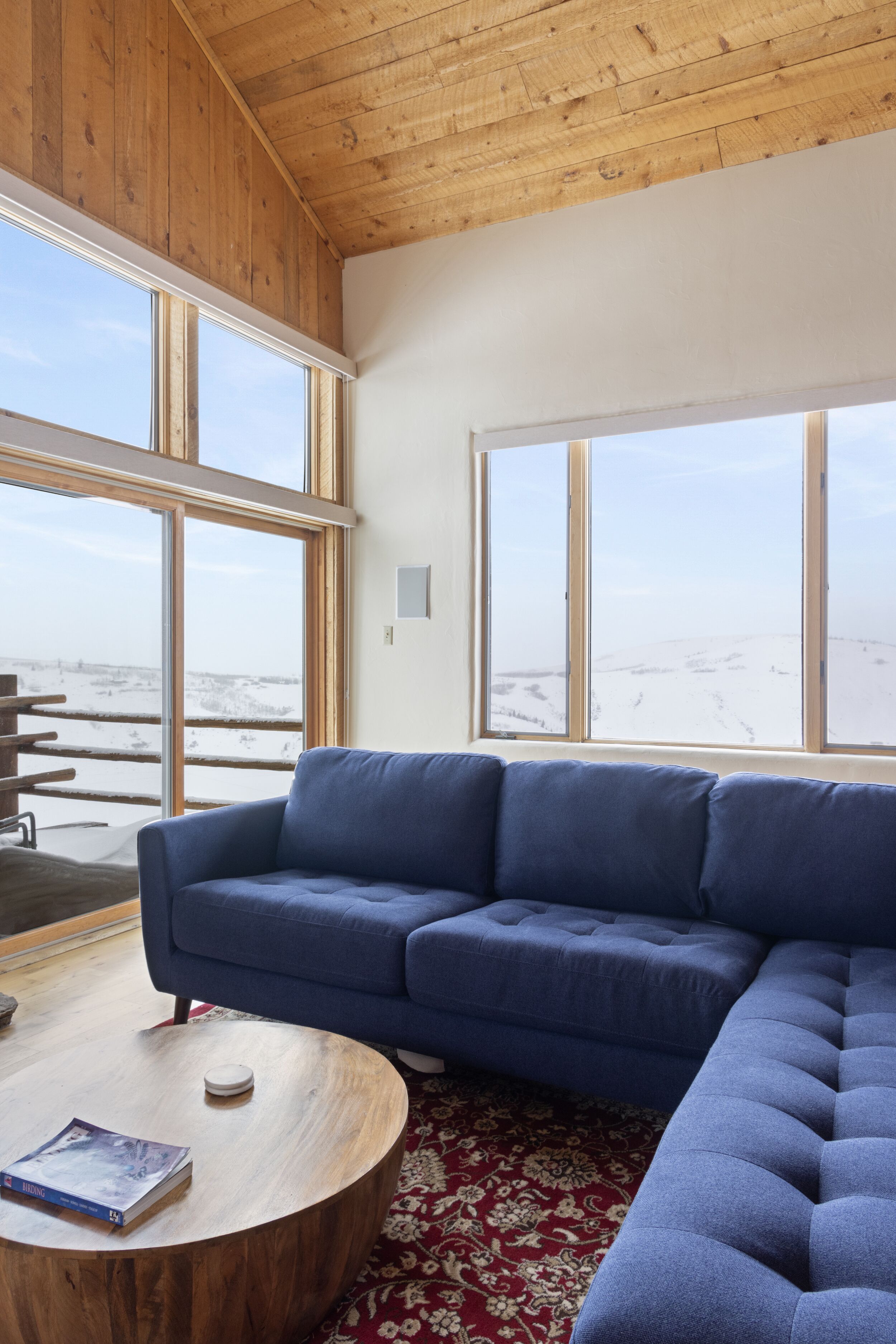 Una sala de estar moderna con un sofá seccional azul, una mesa de centro de madera y grandes ventanas con vistas a un paisaje nevado.