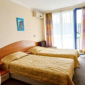 A hotel room with two beds, a nightstand, a painting on the wall, and a window with curtains. - Hotel Pliska (Nessebar)