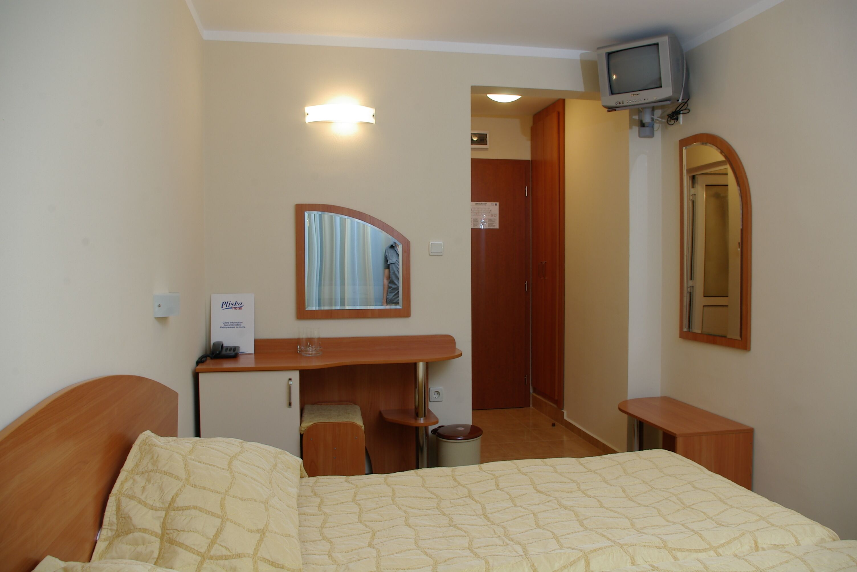 A hotel room with a bed, a desk, a mirror, a television, and a door leading to a hallway.