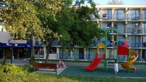 A playground with a slide and climbing structure, surrounded by a grassy area and a building with balconies. - Hotel Pliska (Nessebar)