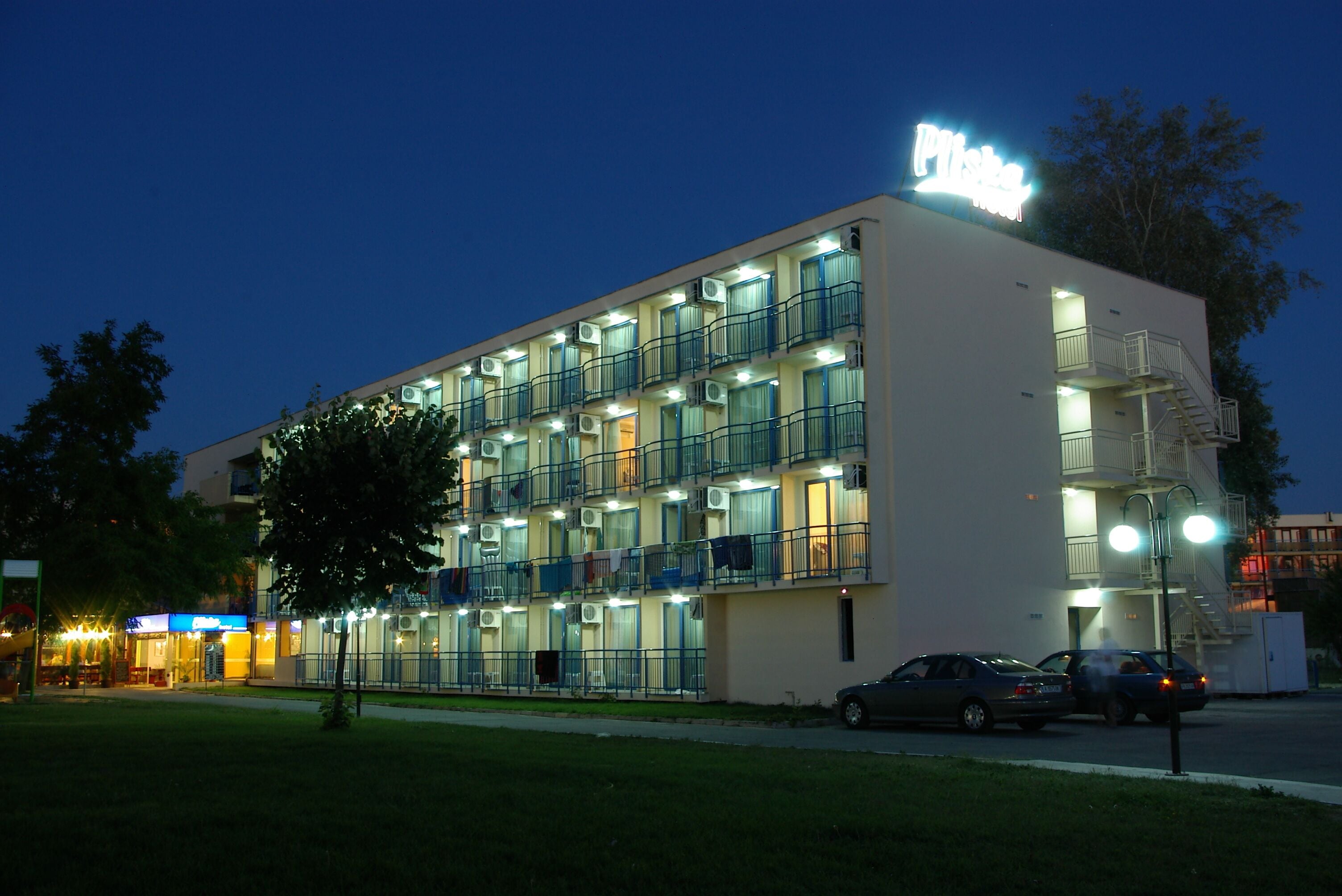 A multi-story hotel with balconies, a visible fire escape, and a parking area with cars.