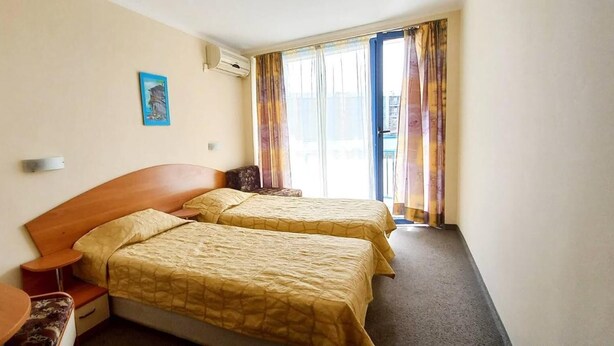 A hotel room with two beds, a wooden nightstand, a small table, and a window with curtains. - Hotel Pliska (Nessebar)