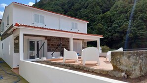 A white building with a red-tiled roof, a covered patio with white chairs, and a stone wall.