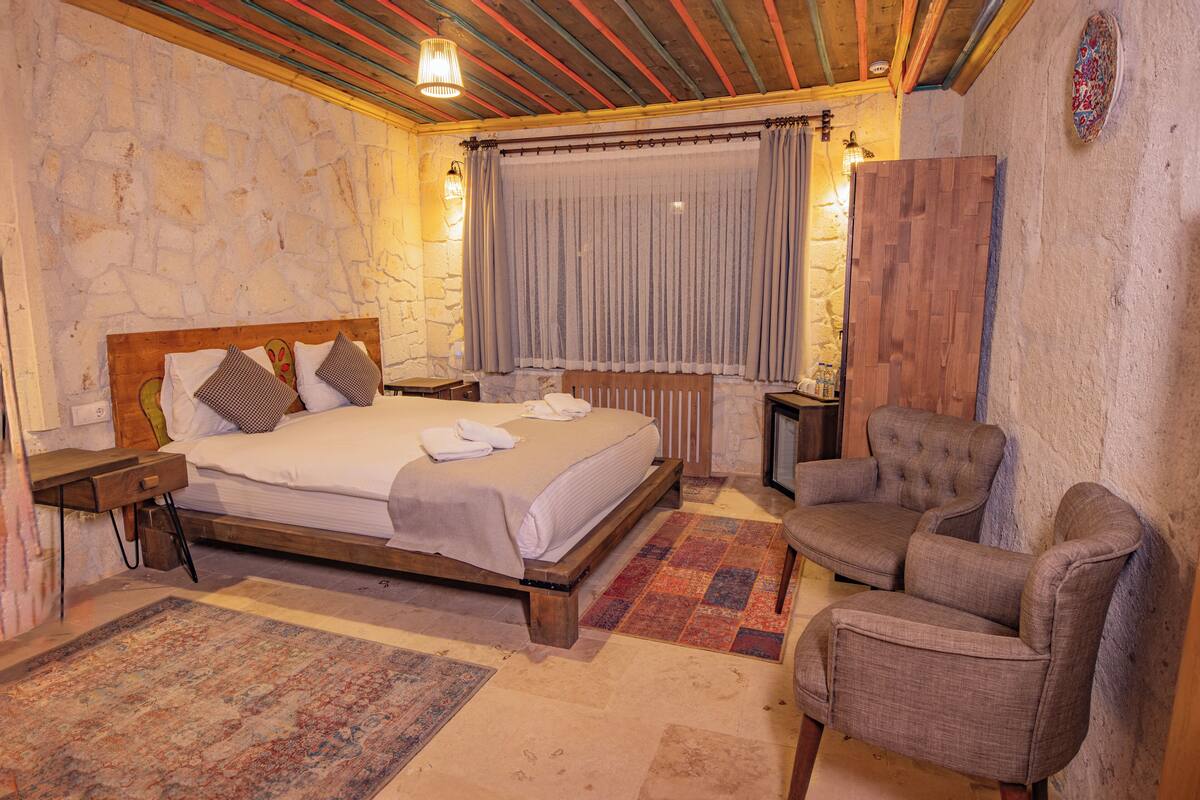 A bedroom with a wooden bed, a chair, a nightstand, a window with curtains, and a stone wall.
