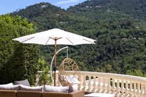 Un canapé en osier avec des coussins, un fauteuil suspendu et un parasol blanc sur un balcon doté d’une balustrade blanche.