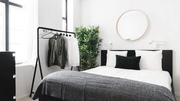 A modern bedroom with a bed, a black and white patterned bedspread, a wall-mounted round mirror, a plant, and a clothing rack with hanging clothes.