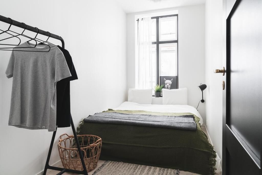 Un dormitorio con cama, perchero, cesta de mimbre y ventana con cortinas.