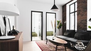 A modern living room with a sofa, coffee table, and a lamp. There is a large window with a view of the outdoors. - Stay Bryggen - Serviced Apartments (Bergen)