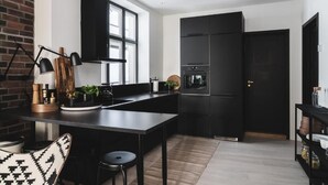 A modern kitchen with black cabinetry, a large window, and a brick wall. - Stay Bryggen - Serviced Apartments (Bergen)
