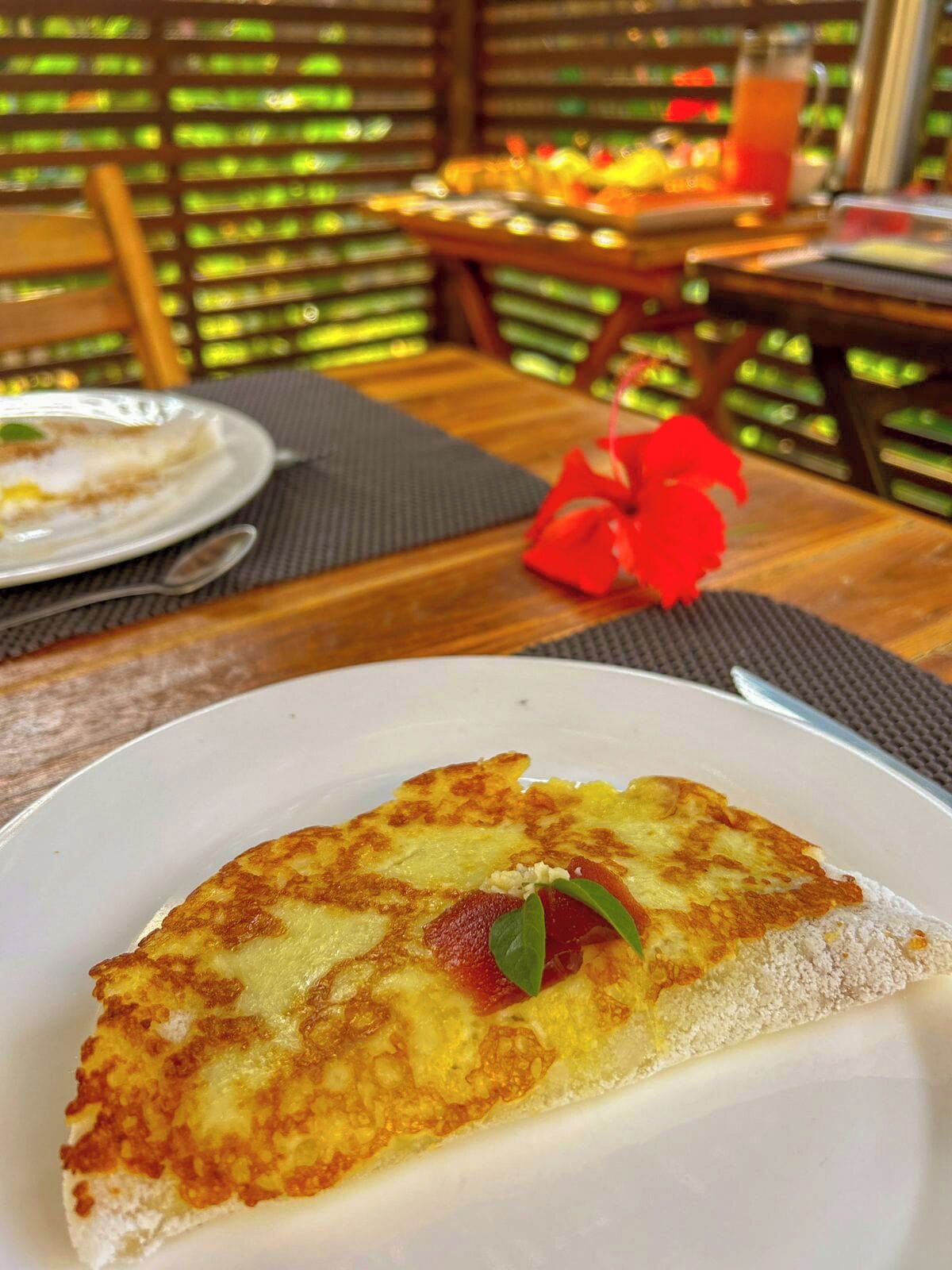 A dining table with a plate of food, a red flower, and a drink in the background.