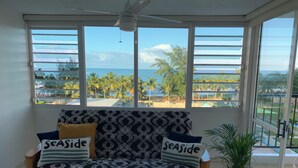 Et værelse med en sofa, der vender ud mod et stort vindue med udsigt over en strand og palmer.