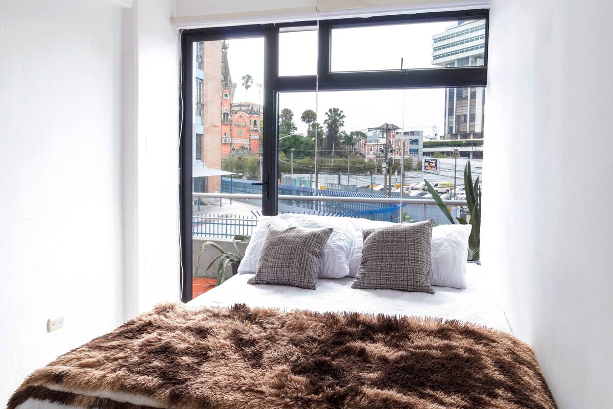 A bedroom with a large window, a bed with a white bedspread and brown fur throw, and a view of a cityscape.
