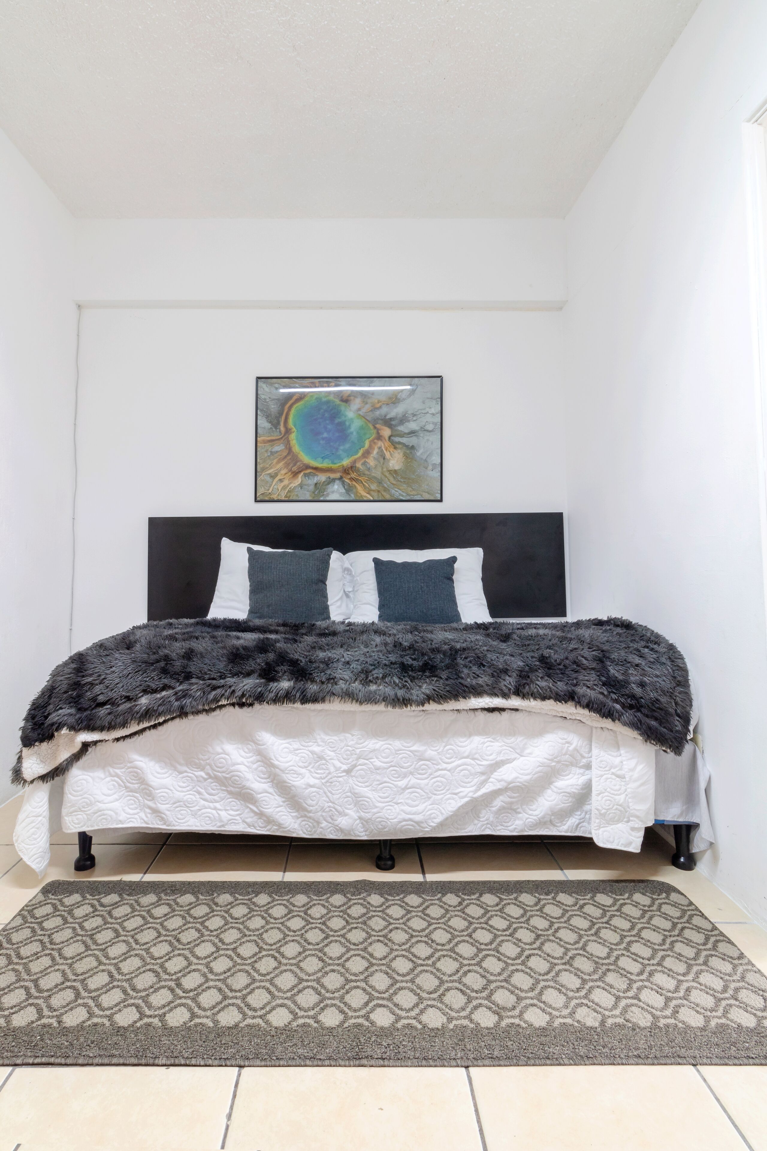 Una cama bien hecha con un cabecero oscuro, un tapete estampado y un cuadro abstracto en la pared.