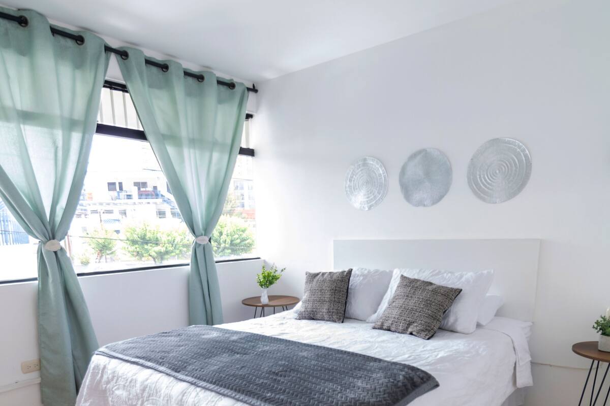 A neatly made bed with a white and gray bedspread, a small bedside table with a plant, and a window with light green curtains.