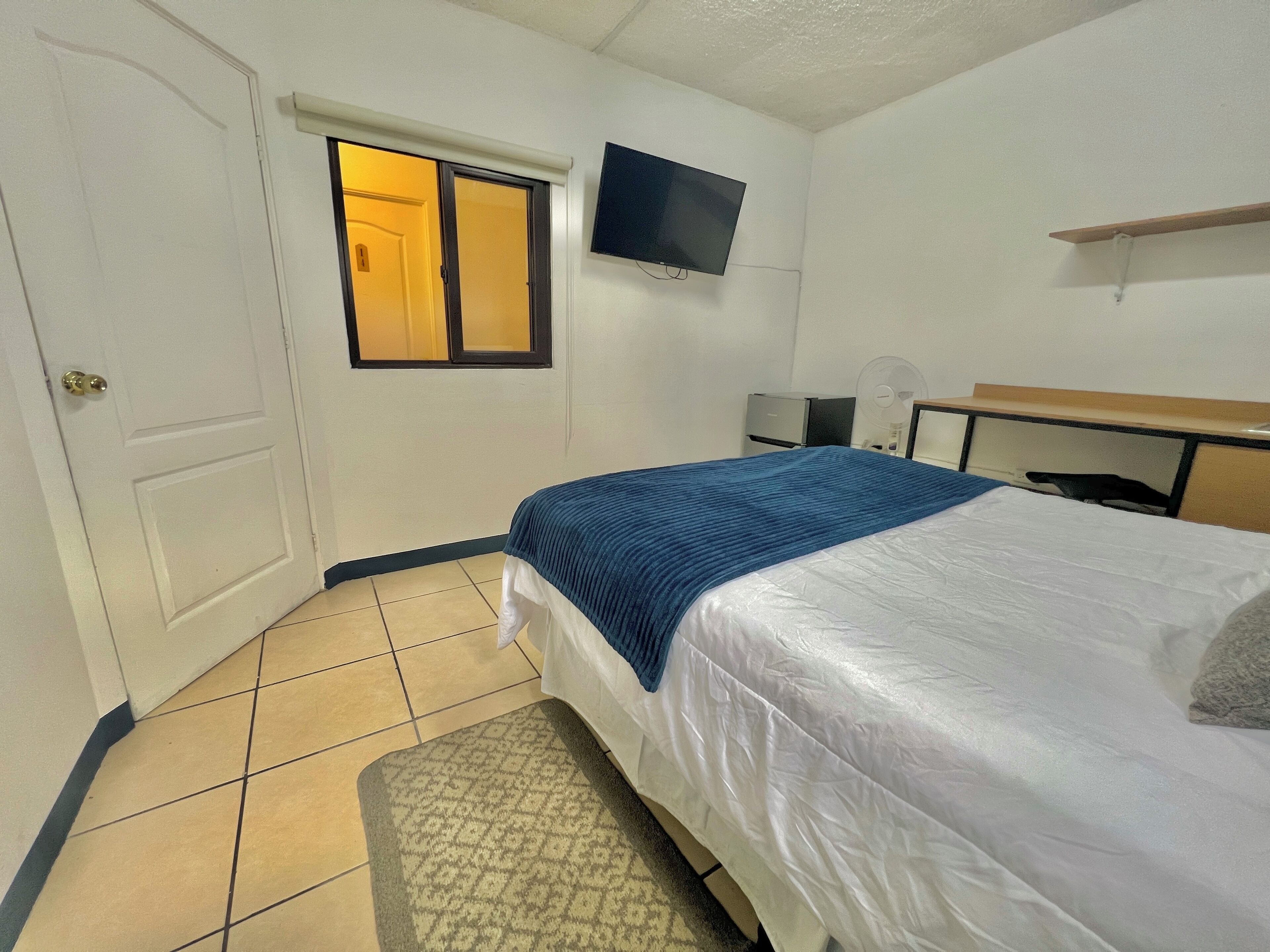A hotel room with a bed, a TV mounted on the wall, a desk with a chair, and a window with blinds.