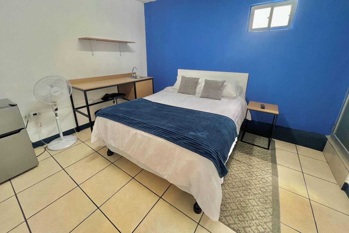 A bedroom with a bed, a desk, a chair, a standing fan, and a small window.